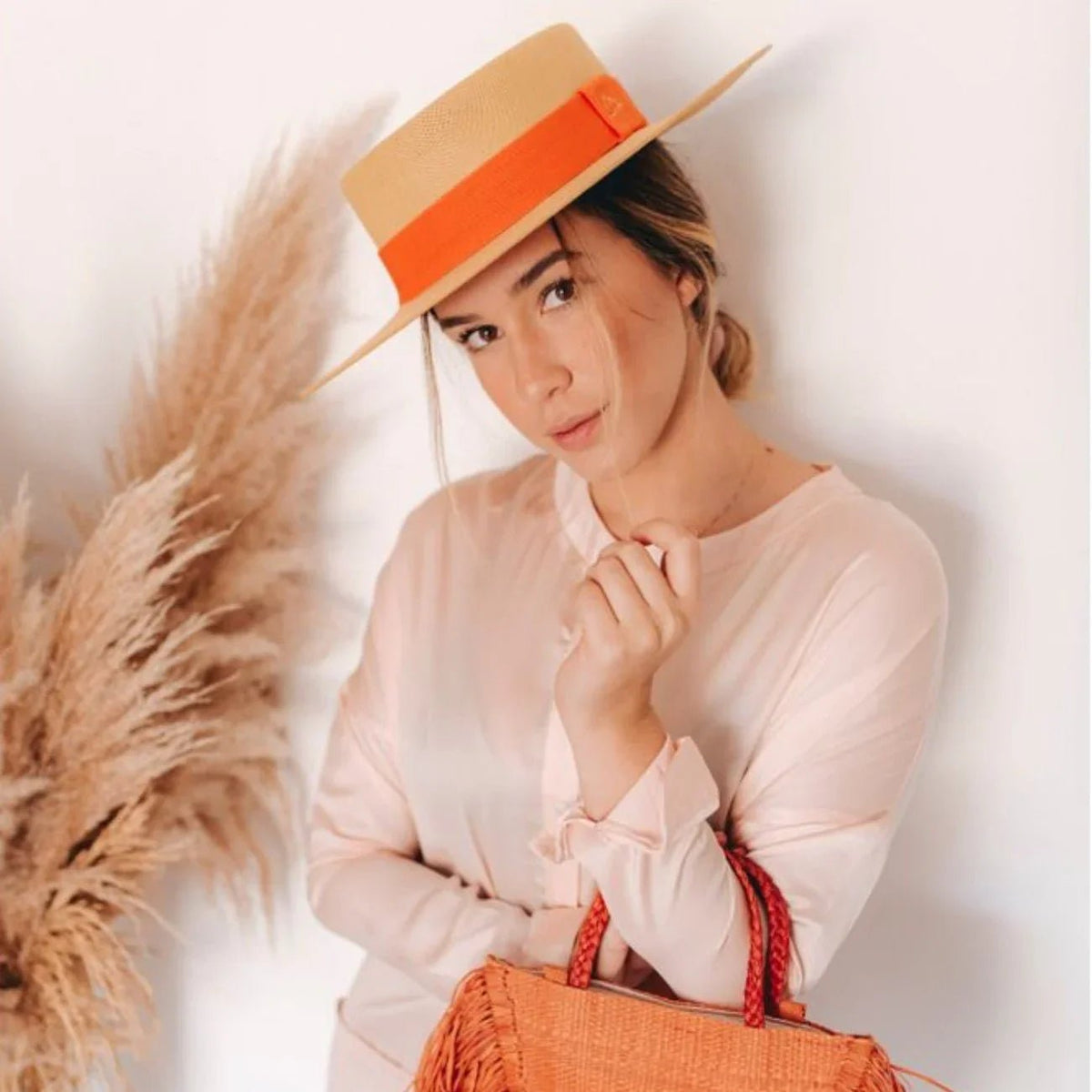 Woman in beige boater hat with orange band, holding woven straw bag, pampas grass background